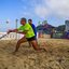 A imagem mostra uma partida de beach tennis (ténis de praia) a decorrer numa faixa de areia à beira-mar, com atletas concentrados no jogo e edifícios urbanos ao fundo