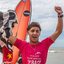 Imagem mostra Kalani Robles, surfista de Ubatuba, comemorando com uma prancha e uma bandeira de São Paulo na faixa de areia da praia