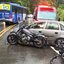 Imagem mostra uma colisão de trânsito em rodovia sob chuva, envolvendo um carro de passeio cinza, duas motocicletas caídas na pista, um ônibus azul ao fundo e uma viatura de resgate