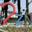 Imagem mostra Telma de Souza em baixo do monumento de coração instagramável com a frase "Eu amo ZNO" localizado na Praça Júlio Dantas, no bairro Caneleira, na Zona Noroeste de Santos