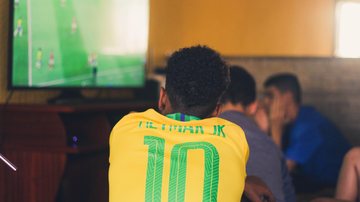 Impactos positivos e negativos da cultura do futebol no Brasil Homem sentado, usando a camisa do Brasil amarela com nome do Neymar, assistindo jogo de futebol - Unsplash